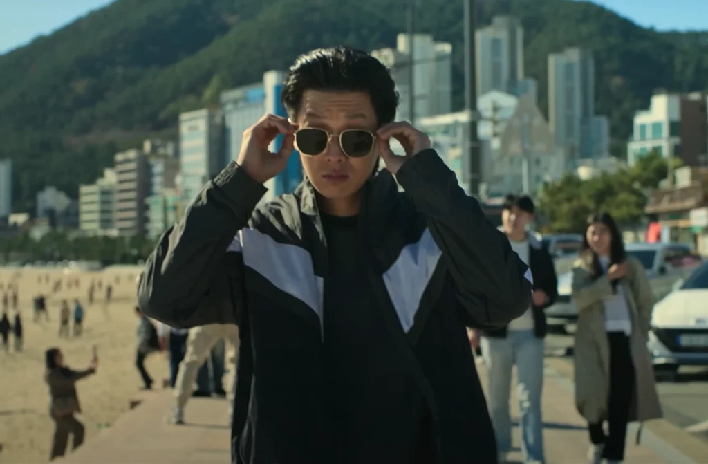 On a sandy beach, a man with dark hair styles his gold-framed sunglasses. He is wearing a black jacket with white accents. In the background, individuals are walking, and buildings can be seen along the beach, which relates to Top 5 Korean Dramas available on Netflix in 2025.