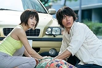 Alice in Borderland: Two individuals, a woman and a man, positioned near a beige vehicle. The woman, with short dark hair, is on her knees, wearing a light green top and grey pants, and appears concerned. The man, sporting medium-length dark hair, is kneeling and leaning towards the front; he is wearing a light-colored button-down shirt, also displaying a worried expression. A figure obscured by a patterned blanket lies on the ground in front of them.