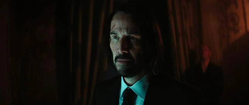 Close-up of Keanu Reeves with a determined expression, slight scarring visible on his forehead, in a dimly lit scene, possibly related to ballerina movie review.
