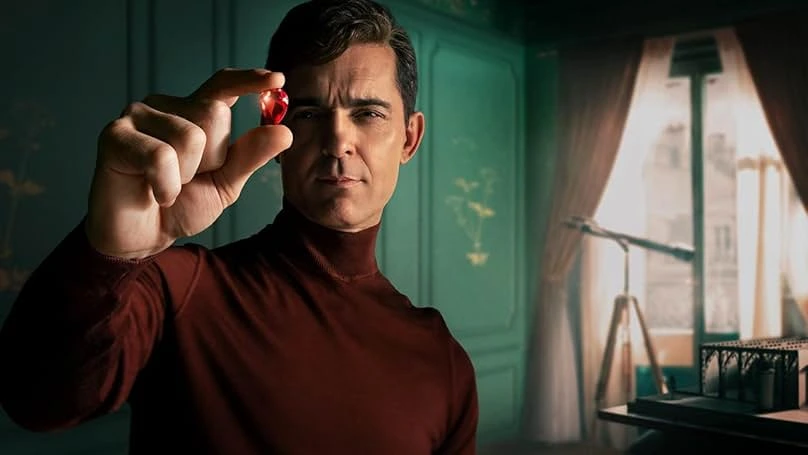A man wearing a maroon turtleneck holding a red gemstone to his eye, with a faintly smug expression, in anticipation of the Berlin Season 2 Release Date. Behind him is a classically decorated room with a table and a scale model of a building, with a telescope in the background.
