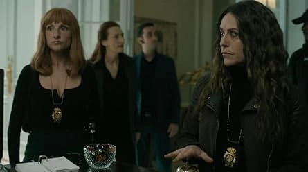 Three people stand in a room; two women in the foreground and a man further back. The woman on the left of the frame has straight auburn hair and wears a black dress and a pendant necklace. The woman in foreground on the right has long, dark, wavy hair and wears a black turtleneck and jacket along with a similar pendant necklace. A man in a black suit is behind the woman on the left, looking to his left. The women appear focused and observant.