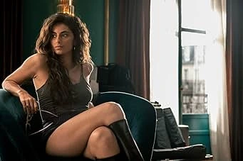 A woman with dark hair and light skin sits angled in an upholstered chair, gazing intently towards the left. She wears a dark top and black boots. Behind her are a green wall and window with bright daylight outside.