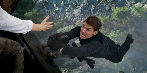 A man in black attire clings precariously to a train mechanism, a distressed expression on his face, with an outstretched hand extending toward him for assistance, set against a blurred mountainous landscape.