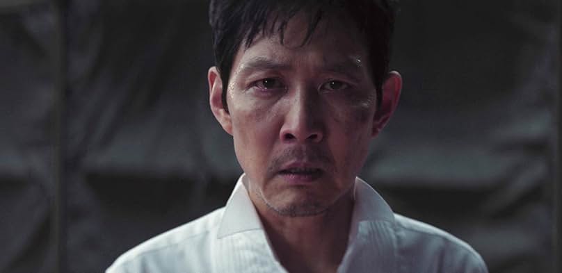 Close-up portrait of Gi-hun from Squid Game Season 3, with visible blemishes and dark circles around his eyes, wearing a white shirt, against a dark background. He appears distressed.