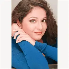 Head and shoulders portrait of Gracy Singh, a fair-skinned woman with long, wavy dark brown hair, wearing a blue sweater and looking directly at the viewer with a neutral expression; Missing bollywood actresses.