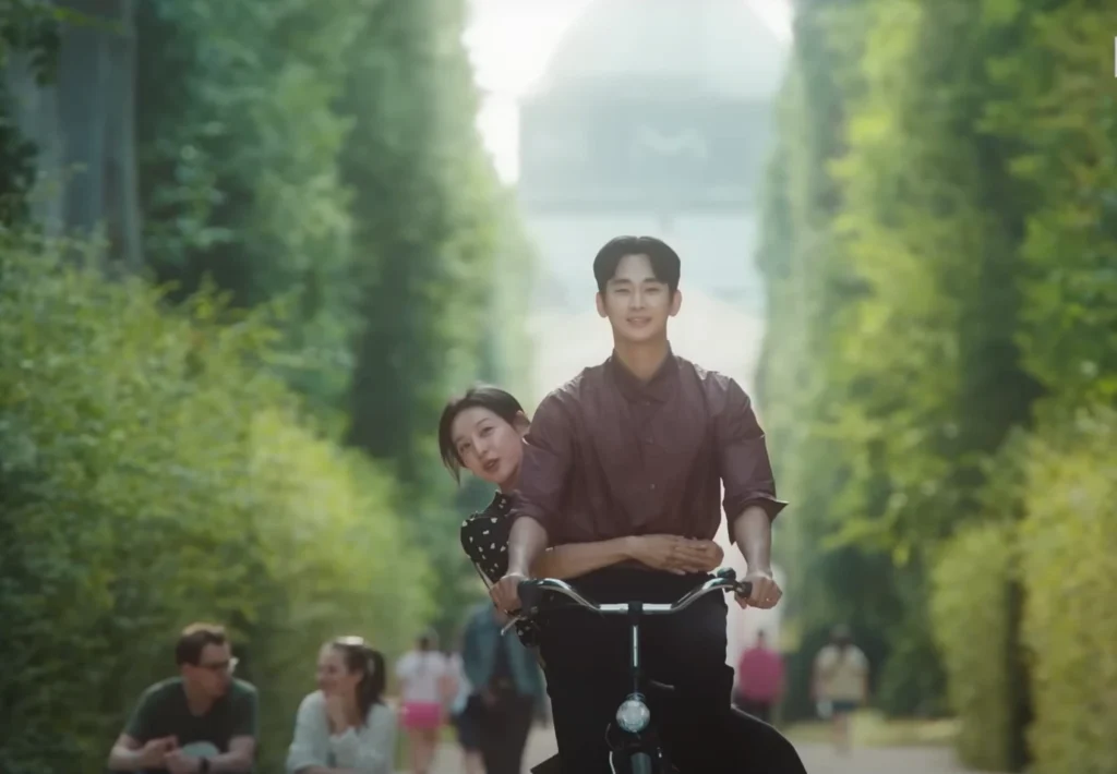 A man and a woman are riding a bicycle on a tree-lined path. The man is in front and smiling. The woman is behind him, also smiling, with her arms around his waist. Part of Top 5 Korean Dramas available on Netflix in 2025.