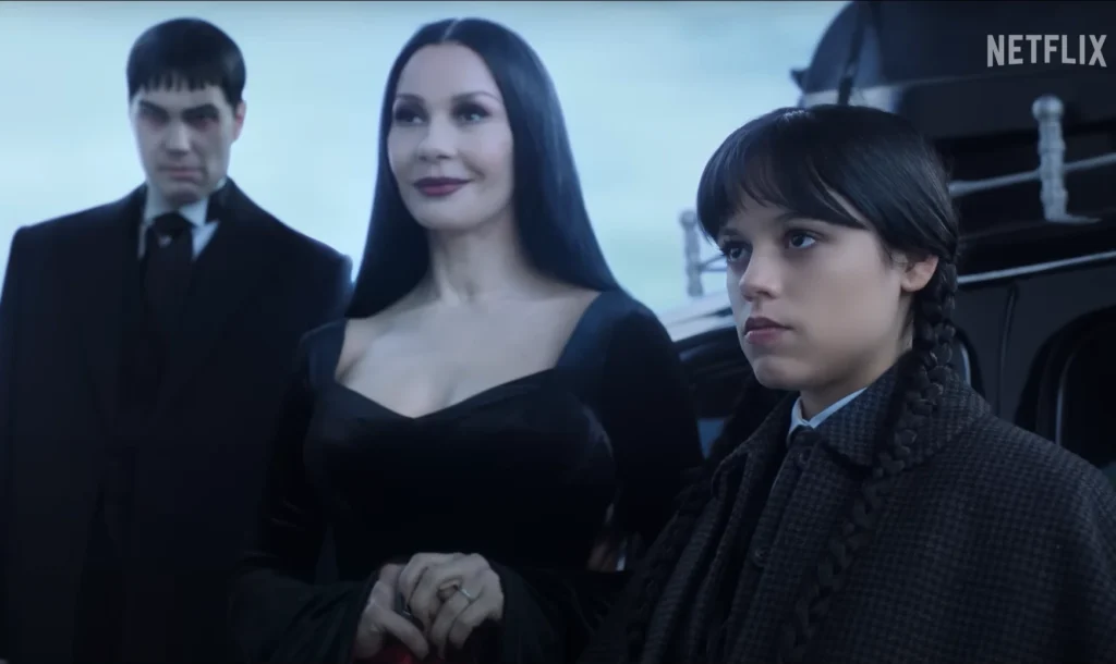 Still from the Netflix television series "Wednesday Season 2 Release Date," featuring characters Gomez, Morticia, and Wednesday Addams. Gomez, dressed in a black suit, stands to the left with a neutral expression. Next to him is Morticia, wearing a black velvet dress and smiling subtly. Wednesday stands on the right, in a dark tweed coat with two braids, and looking intently at something unseen. The Netflix logo appears in the upper right corner.
