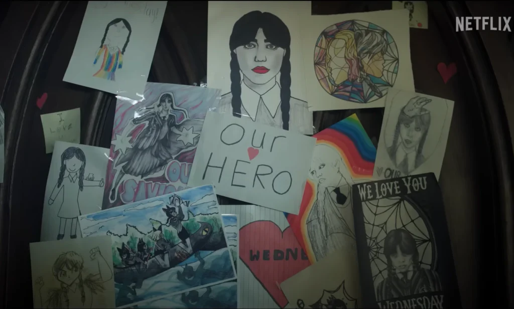 Close-up shot of artwork depicting Wednesday Addams; drawings and posters, some with written messages such as "Our Hero" and "We Love You," are affixed to a wooden surface, conveying admiration and support for the character.