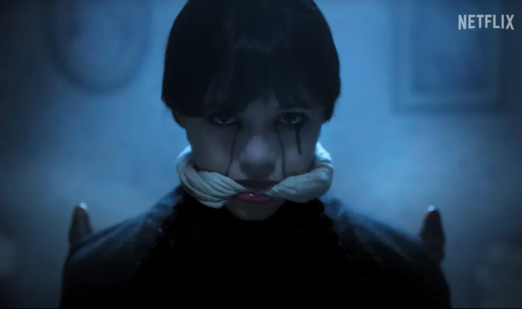 Close-up of Wednesday Addams with a neutral expression, black hair, and a white gag around her mouth, with streaks of black paint extending downward from her eyes.
