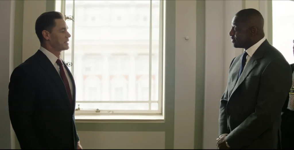 In Heads of State reviewing, John Cena and Idris Elba in suits converse near a window, Cena with a slight smile.