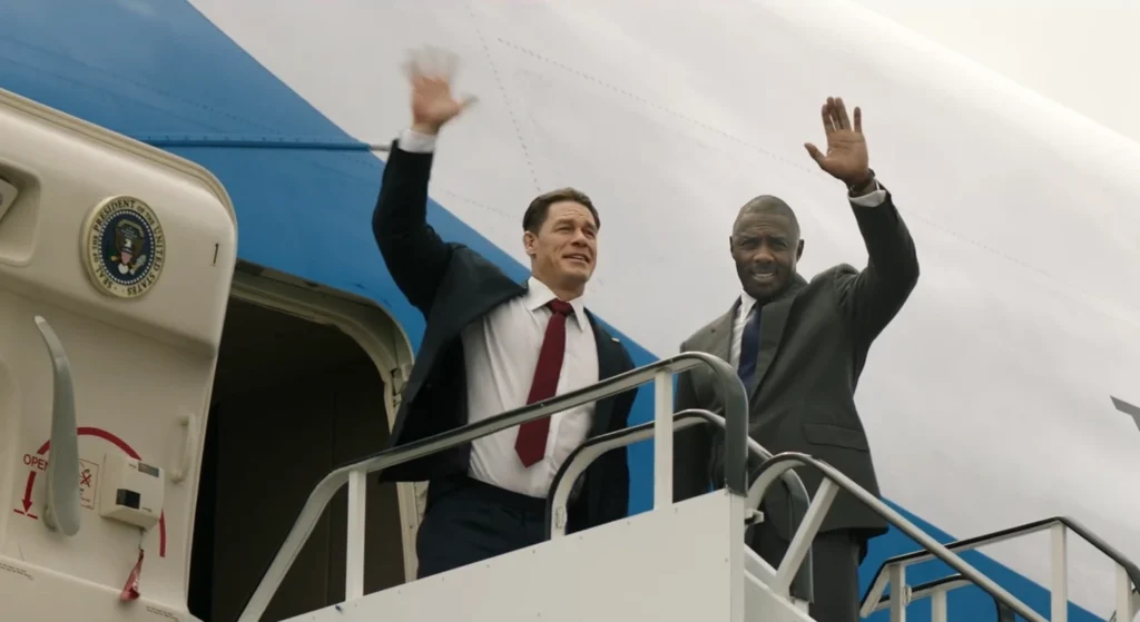 From Heads of State, John Cena and Idris Elba stand on the steps of Air Force One, waving. John Cena is smiling widely and wears a black suit with a red necktie, while Idris Elba, in a charcoal suit, offers a more reserved smile. The Presidential Seal is visible on the aircraft.