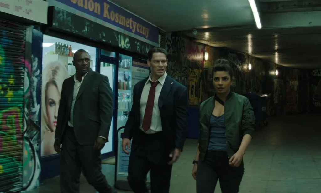 A medium shot shows Idris Elba, John Cena, and Priyanka Chopra Jonas walking down a dimly lit corridor with posters on the wall. Elba, on the left, wears a grey suit and his expression is serious. Cena, in the center, wears a dark suit with and a red tie, looking forward. Chopra Jones, to the right, wears a green jacket. All three appear to be headed in the same direction.