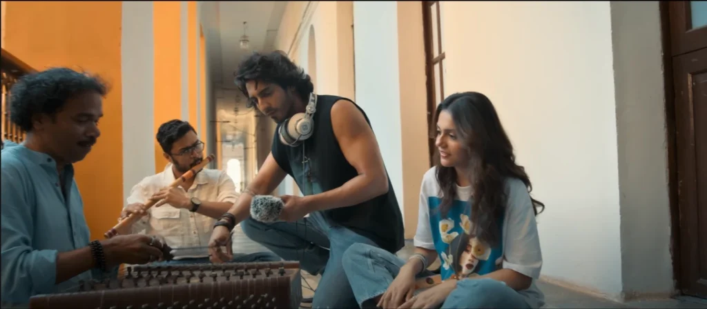 Scene from Saiyaara Movie Review: A musical ensemble comprising four individuals, a woman with a subtle smile on her face, positioned to the left of the frame, sits in close proximity to a man, his gaze focused downward as he handles a microphone; to their left, two men perform on instruments, one playing a flute, the other a stringed instrument; the musicians are located indoors, along a hallway with decorative pillars.
