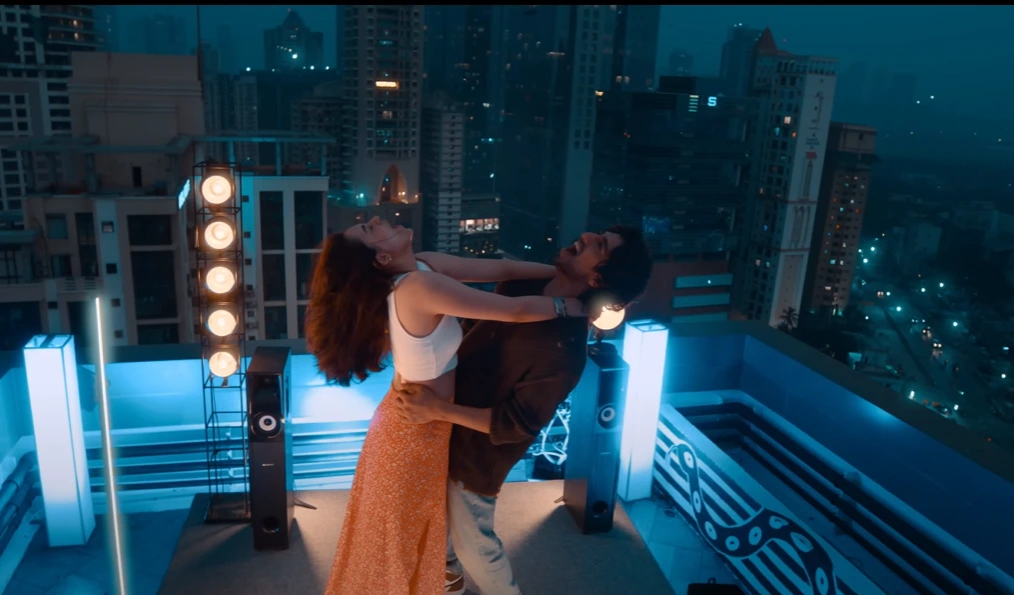 On a rooftop, a man and woman embracing, with their heads tilted back and laughing expressions, against a backdrop of city skyscrapers at dusk. Decorative lighting fixtures add to the rooftop ambiance. Saiyaara box office collection day 10.