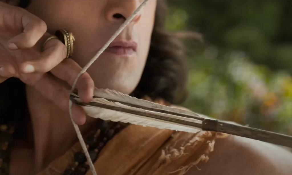 Close-up shot of actor Ranveer Kapoor, as Lord Ram, preparing to aim an arrow in Ramayan Movie Namit Malhotra production. The arrow's feathered fletching is in focus, drawn back against the taut bowstring. The man's expression is serious. His fingers hold the bowstring taut and sport jeweled rings.