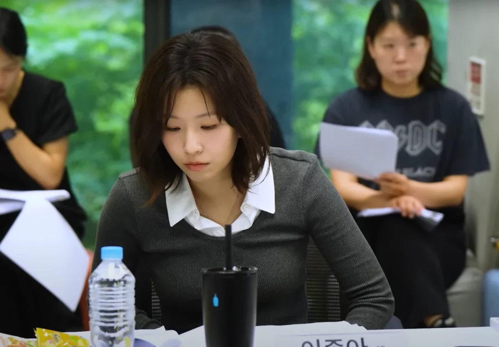 At a table read for All of Us Are Dead Season 2, a young Asian woman is looking down at a script. Her expression is thoughtful. Another woman in the background wears an AC/DC t-shirt.