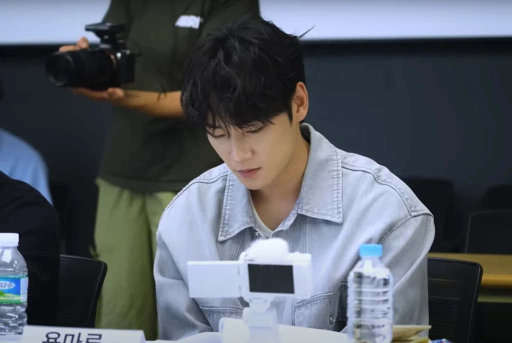 Actor as part of the "All of Us Are Dead season 2" cast, sits at a table with papers. He is fair-skinned, and has short black hair He is wearing a light-blue denim jacket. His head is bowed down, and he is calmly reading the papers. To the left is a person holding a professional camera. In front of him are a white personal recording camera and a bottle of water.