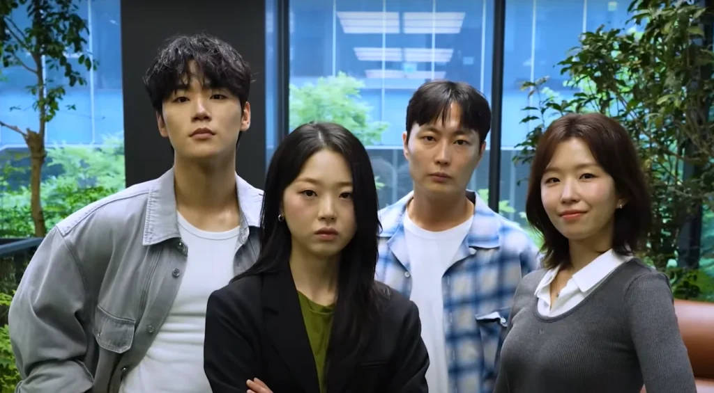 Four cast members for All of Us Are Dead Season 2 stand in a row with flat expressions. The members are of Korean descent. The two center members are slightly closer to the camera; the member second to the right smiles slightly. A green atrium is visible in the background, with blue panes behind that.