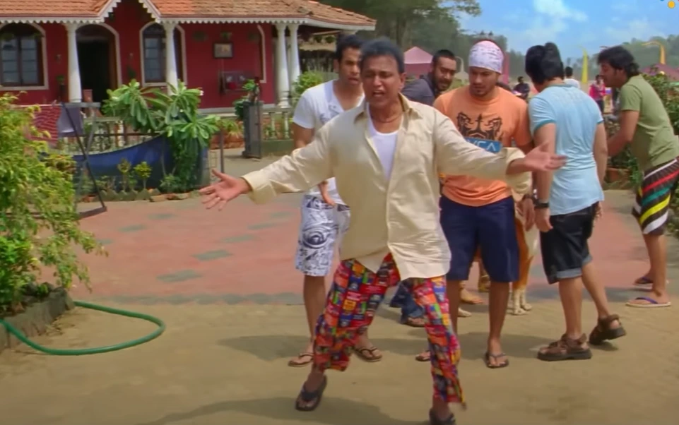 A medium shot depicts a man in the foreground with open arms and mouth ajar, conveying shock or surprise. He wears an unbuttoned beige shirt over a white tee and colorful pants. Behind him, several men look on with expressions ranging from amusement to confusion. The scene appears to be outdoors, possibly a compound with a red-roofed building visible to the left. This setting suggests a moment captured during the filming of "Golmaal 5."