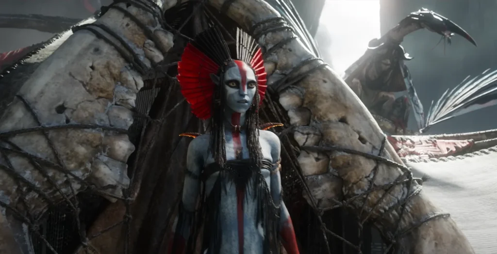 Character from Avatar 3 trailer with light blue skin, red body paint, and a red feathered headpiece, standing in front of a structure made of bones. The character wears a serious expression.