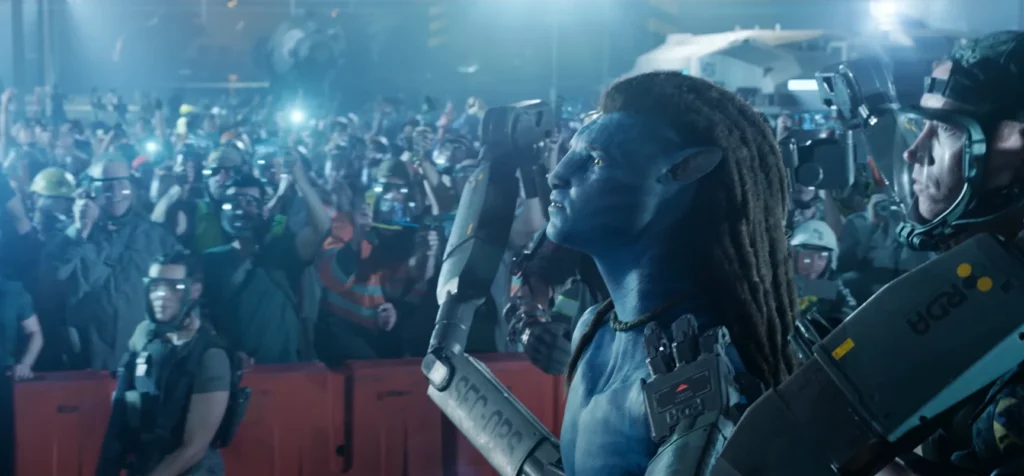 An Avatar 3 trailer scene with a crowd of humans appearing happy, some holding up lights. To the right is a serious-looking man wearing a helmet and holding equipment labeled "RDA." In the foreground, an Avatar character with blue skin and dreadlocks is visible from the chest up, also looking serious. The scene suggests anticipation or celebration.