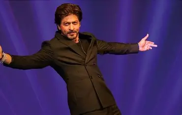 Close-up of Shah Rukh Khan, in mid-motion, facing the viewer and smiling, against a gradient purplish-blue background. Shah Rukh Khan is wearing a black suit.