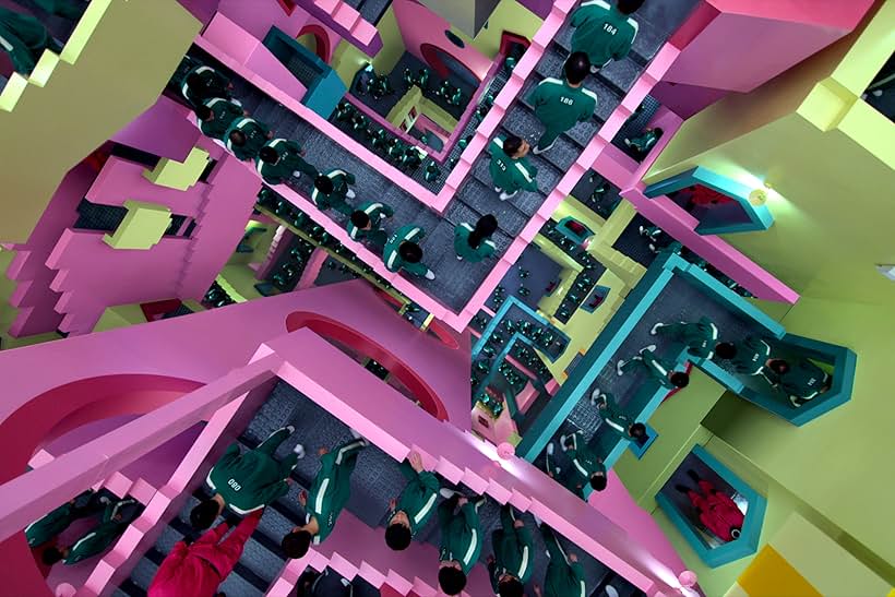Looking down at a maze of vibrantly colored stairs and landings in shades of pink, green, and blue, numerous figures wearing green tracksuits with white number are walking on the stairwells.