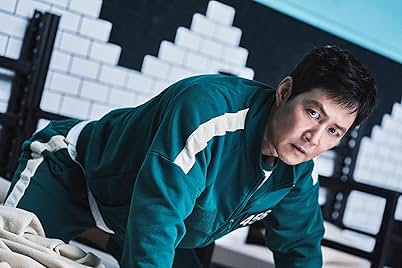 Gi-hun from Squid Game, wearing a teal tracksuit with white stripes, gazes intently forward. The background features a wall with symmetrical white blocks.