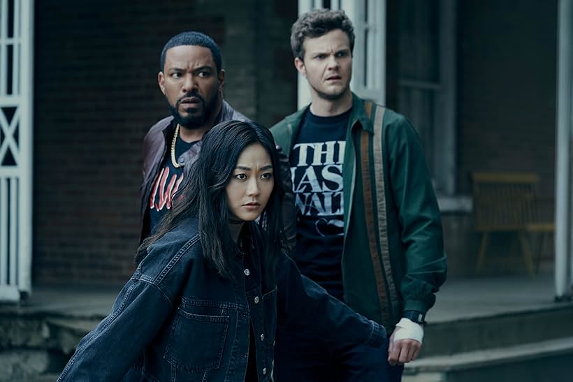 Three individuals stand with serious expressions outside a brick building. The woman in front, with long dark hair and wearing a dark denim jacket, looks slightly downward; the two men behind gaze forward with concern.