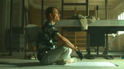 Man wearing a floral shirt and work gloves sitting cross-legged on the floor with a serene expression. Construction materials are stacked behind him.