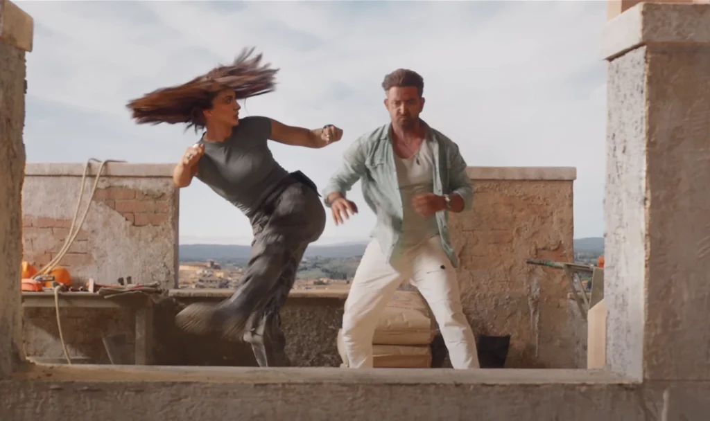 From the "War 2" trailer, an outdoor shot features a woman with flowing hair forcefully kicking towards a man, both exhibiting tense expressions, set against a partially constructed backdrop.