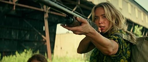 Still from "A Quiet Place Part III" depicts Evelyn, played by Emily Blunt, holding a shotgun with a determined expression. She's wearing a green floral shirt and has a backpack on. Brown metal beams and a green wall are in the background.