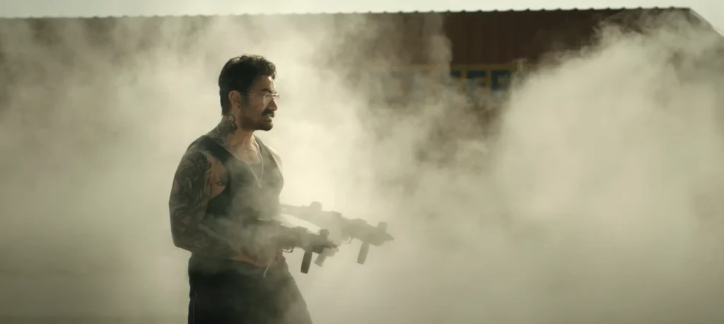 Amir Khan in a tank top and tattoos, holding two machine guns, stands determinedly amidst a fog, likely extracted from the Coolie Trailer Rajnikanth footage.