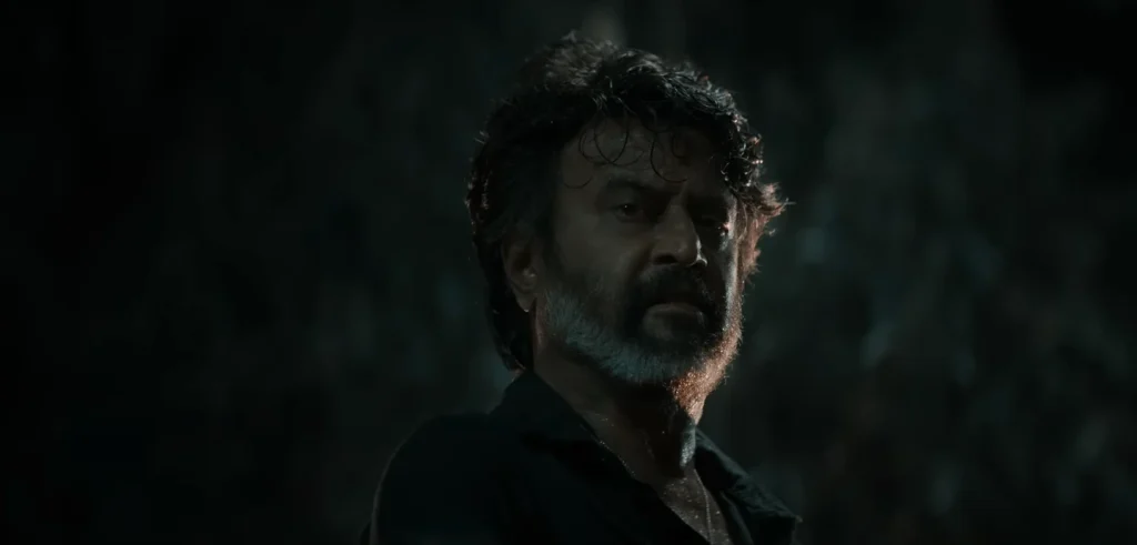 A close-up shot from the Coolie Trailer features Rajnikanth, with a somber expression, set against a dimly lit backdrop. The overall mood is intense and dramatic, consistent with a serious scene from the trailer.