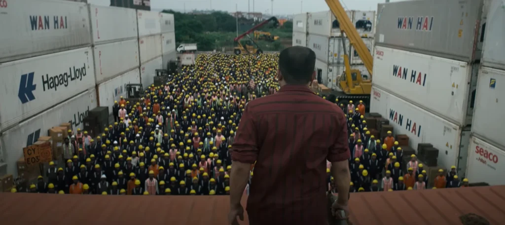 Coolie Trailer Rajnikanth: Rear view of a man in a short-sleeved maroon shirt overlooking a large crowd of laborers in yellow hard hats, with rows of shipping containers and cranes visible in the background.