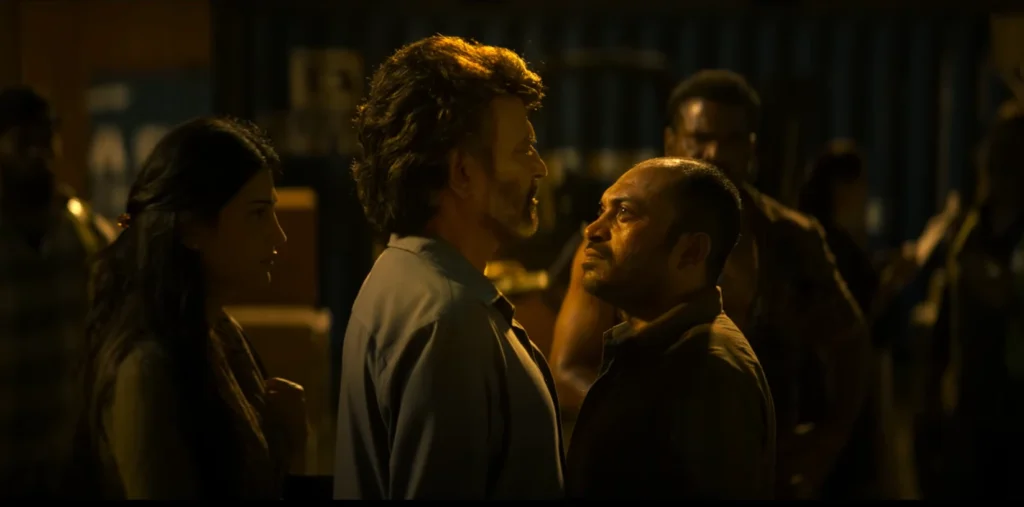 From the Coolie Trailer, Rajinikanth is shown in a medium shot appearing to confront a man wearing a dark shirt, both exhibit serious facial expressions; a woman stands to Rajnikanth's left, looking concerned. The background features numerous shadowy figures with stern expressions, obscuring the details of the setting.