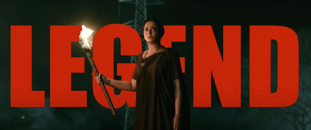 "Ghaati Trailer" still: a woman holds a burning torch in front of a towering metal construction, partially obscured by the large, red, sans-serif letters that spell out "LEGEND". The woman wears a somber expression and is dressed in simple, dark clothing.