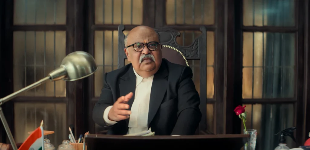 Still from the Jolly LLB 3 teaser featuring Saurabh Shukla as Justice Surendralal Tripathi, wearing spectacles with a white mustache, seated at a desk with an Indian flag and a single red rose.