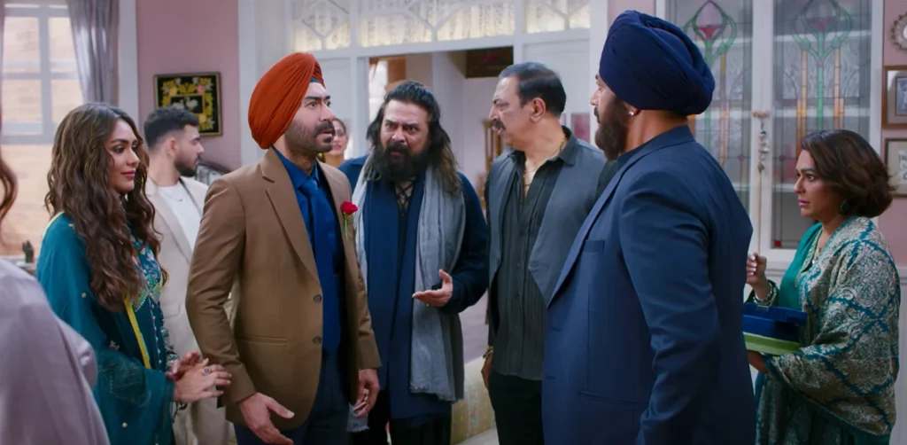 A still from the Son of Sardaar 2 review, featuring a group of concerned-looking people in an ornate room. Ajay Devgan, wearing a tan blazer and an orange turban with a red rose on his lapel, looks off-camera with a worried expression. Surrounding him are other actors with similarly concerned expressions, some wearing turbans and traditional Indian clothing.