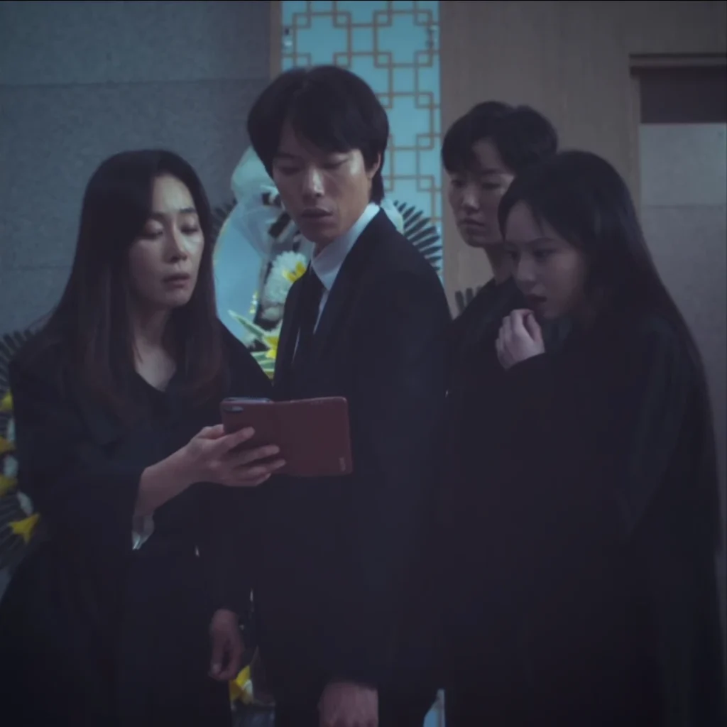 Scene from "The 8 Show" depicts four solemn individuals in dark attire, presumably at a funeral; a woman holds a tablet, exhibiting a concerned expression, while a man observes the screen with furrowed brows.
