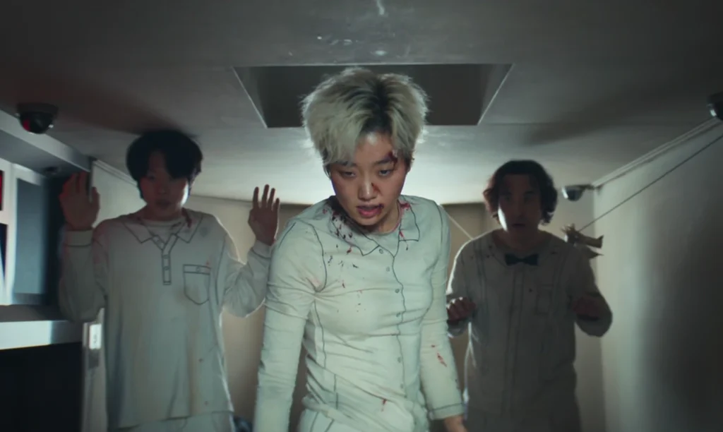 Scene from The 8 Show depicting three individuals in matching off-white pajamas with drawn-on details, appearing distressed with some bloodstains, walking within a confined corridor monitored by surveillance cameras.