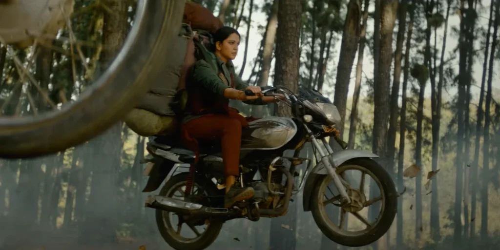 "Ghaati Trailer" features a woman with a serious expression riding a motorcycle through a forest. A second motorcycle wheel is visible mid-air in the foreground.