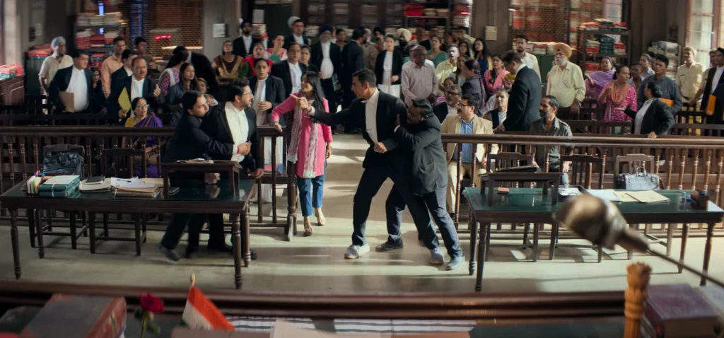 Captured from the Jolly LLB 3 teaser, the scene depicts a crowded courtroom with heightened activity; in the foreground, Akshay Kumar in a black suit extends an arm towards an individual while appearing to restrain another, and various courtroom attendees observe the action with mixed expressions.