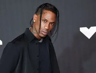 A medium shot of rapper Travis Scott posing against a dark background. He is wearing a black collared shirt and a silver chain, sporting his signature braids. Travis Scott in The Odyssey