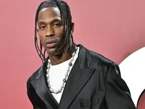 A portrait of Travis Scott wearing a black satin blazer over a white t-shirt and a chunky silver bead necklace. He is posed against a soft pink and white studio background.