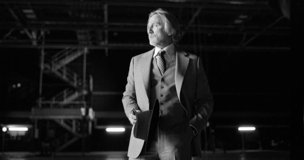 Black and white film still of Daniel Craig as detective Benoit Blanc in a three-piece suit holding a fedora hat in "Wake Up Dead Man."