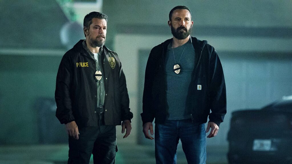 Film still from "The Rip" showing Matt Damon and Ben Affleck in dark jackets with police badges hanging from their necks, standing in a dimly lit industrial setting.